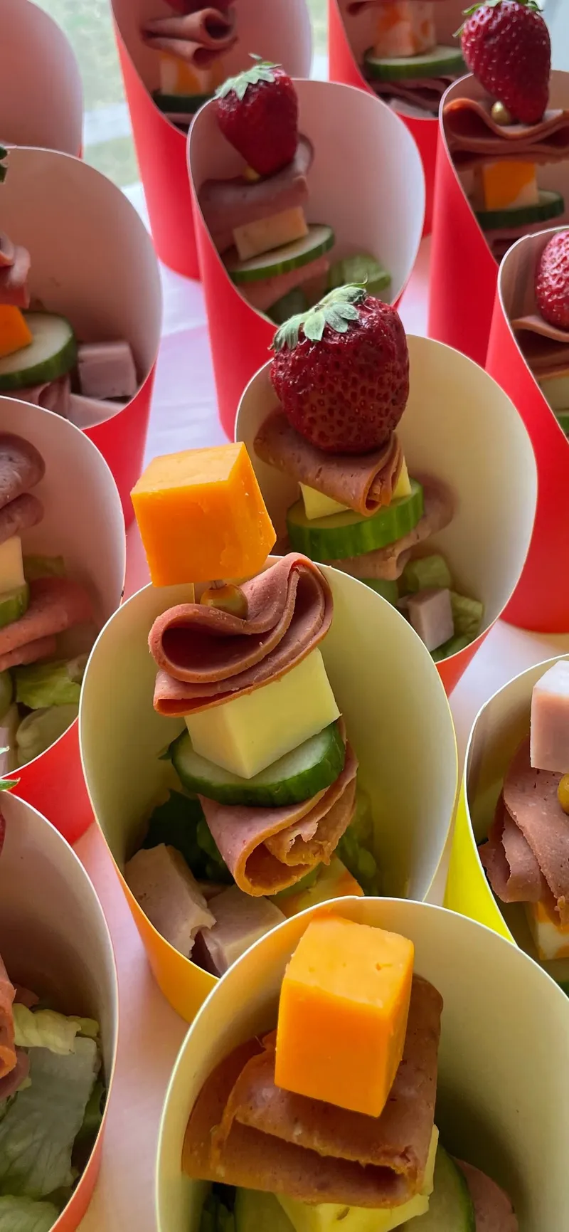 Paper cones holding skewers of ham, cheese, cucumber and strawberry